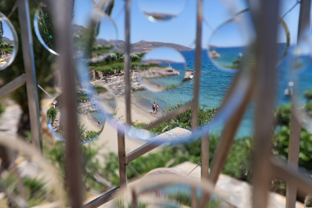 View of a beach with people wading in the water, framed by reflections in a decorative metal structure.