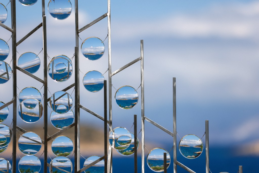A close-up view of a metallic sculpture featuring multiple circular glass elements reflecting a scenic ocean landscape against a blue sky.