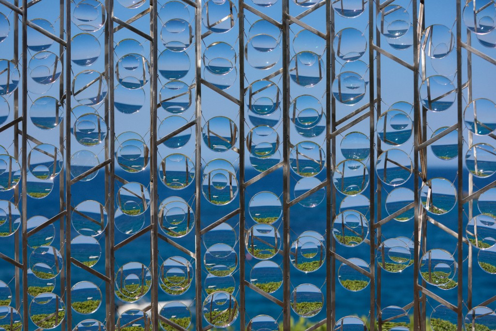 A lens-like structure creates reflections of the ocean and greenery behind it, set against a clear blue sky.