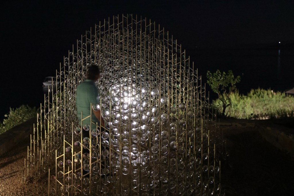 A person standing beside a large artistic installation made of glass spheres and metal rods, illuminated at night, with a scenic background of water and vegetation.