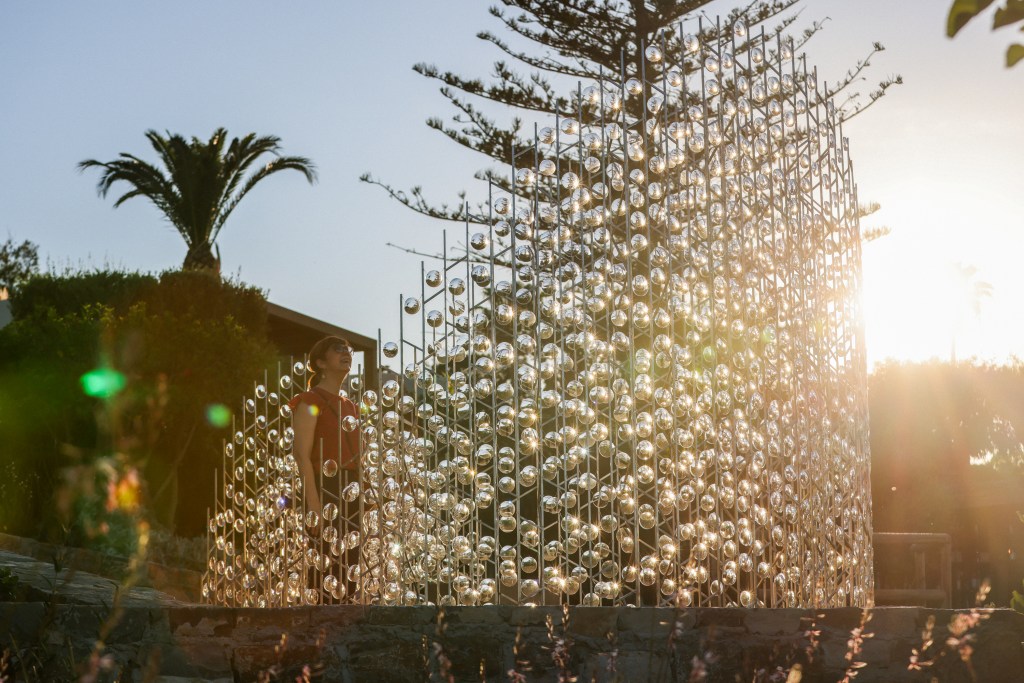 A person standing near a large installation made of transparent orbs set against a sunset background, surrounded by greenery and palm trees.