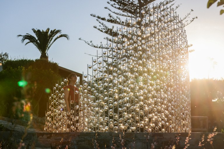 A person standing near a large installation made of transparent orbs set against a sunset background, surrounded by greenery and palm trees.