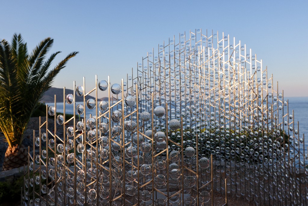 Art installation featuring metallic structures and glass spheres, set against a coastal background at sunset.