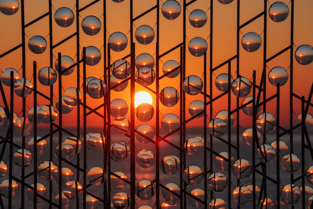 A sunset viewed through a series of reflective spheres arranged on a metal structure, creating an artistic display against a vibrant orange and blue sky.