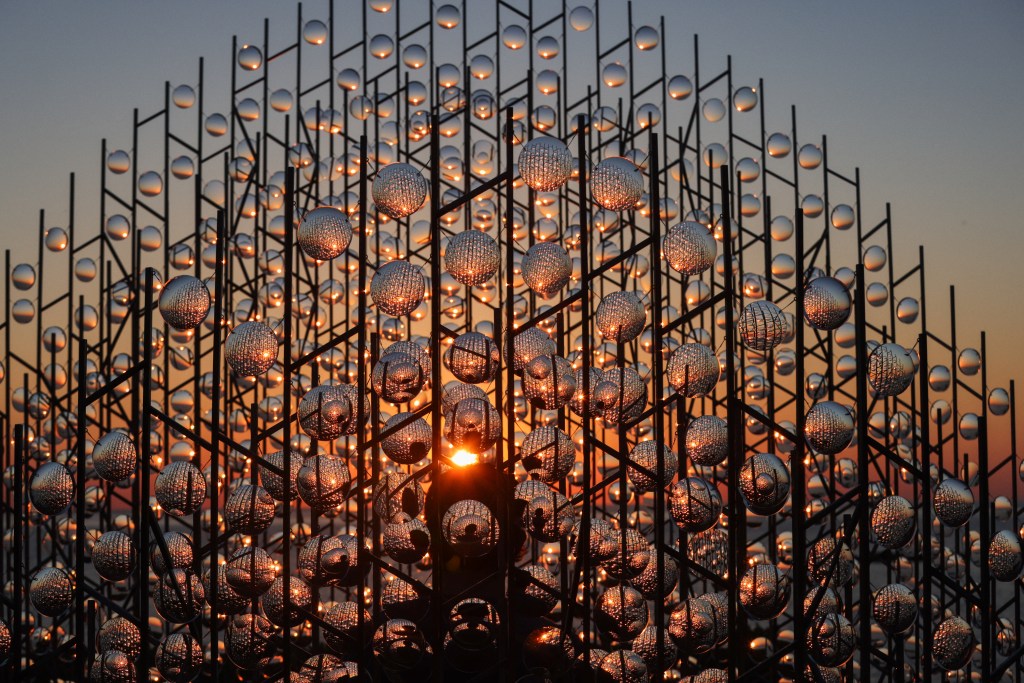 A striking sculpture made of reflective spheres arranged on vertical rods, illuminated by the setting sun, creating a warm glow against a colorful sky.
