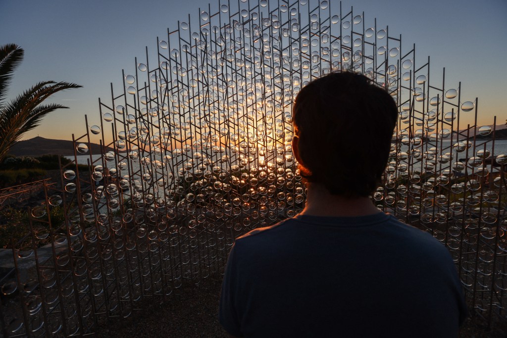 A person standing in front of a large sculpture made of metallic rods and glass bubbles, viewing a sunset over the sea.