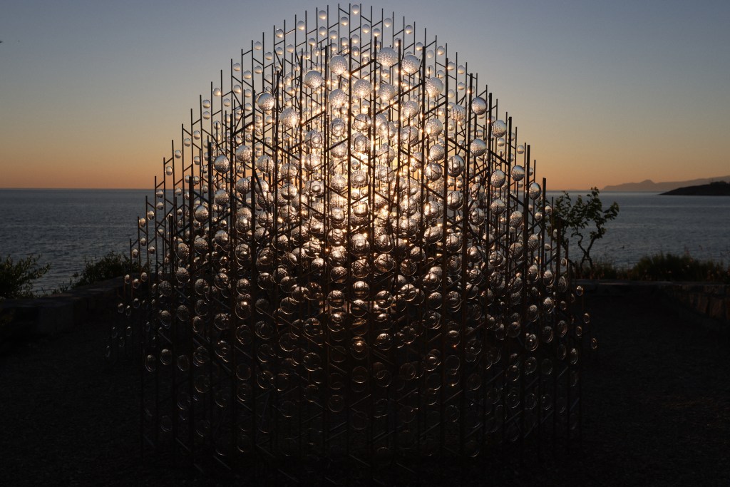 Art installation made of glass discs and metal rods, silhouetted against a sunset over the ocean.