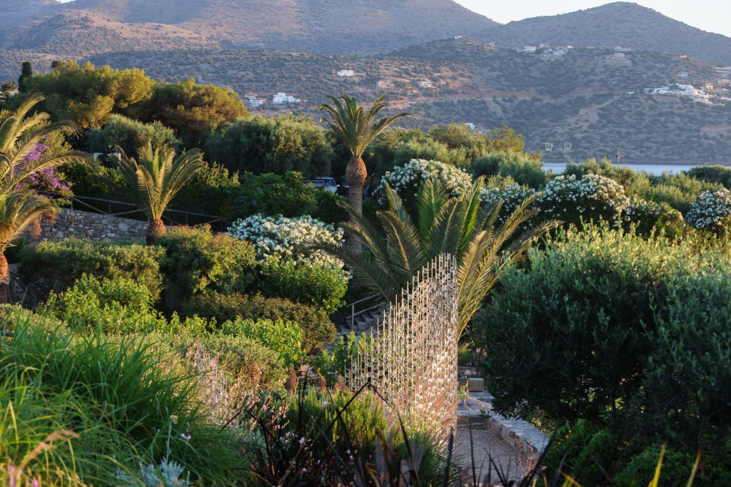 A lush garden scene featuring palm trees and flowering bushes, with a backdrop of mountains and a calm sea in the distance, illuminated by soft evening light.