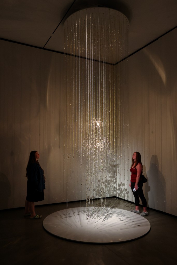Two individuals admiring a hanging light installation with strands of crystal-like elements creating reflections and shadows on the floor.