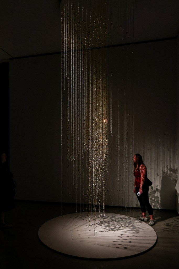 A person observing an art installation featuring hanging strands of light reflecting off a circular base, creating intricate shadows on the ground.