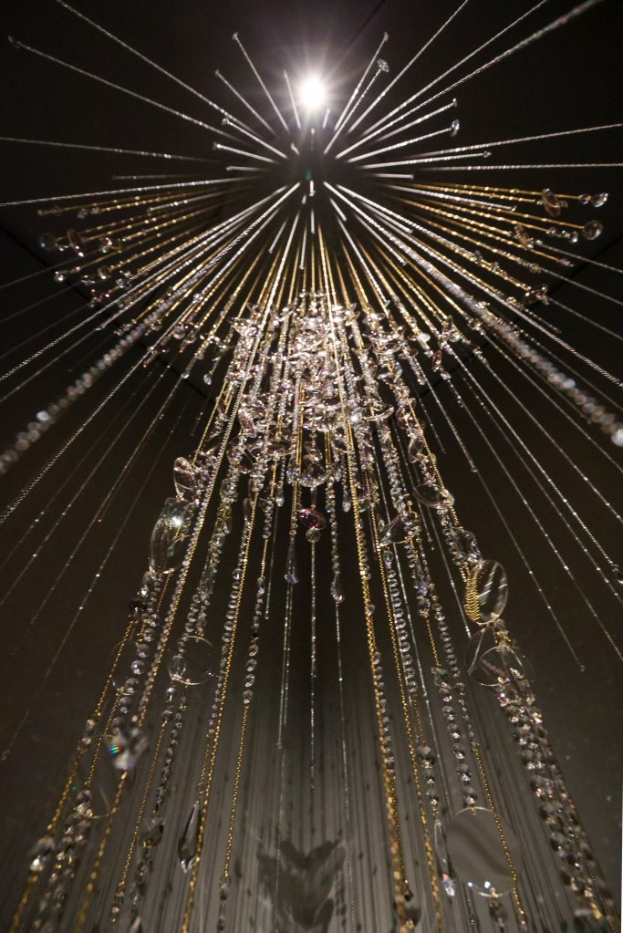A stunning chandelier installation viewed from below, featuring shimmering crystals and gold strands radiating upwards towards a central light source.
