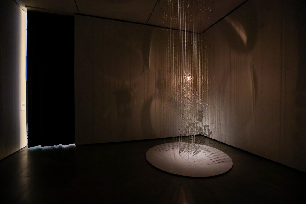 An art installation featuring suspended strands of crystals casting light and shadows in a dimly lit room, with a circular base beneath.